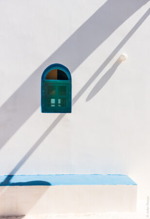 The shadow of an arch supporting a bell falls across the side of the small church of Aghios Petros. (Jun18)