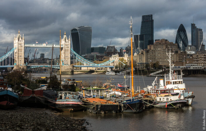 Tugs and barges moored in dramatic light on the south side of the Thames with Tower Bridge also in the light but City towers on the north side under dark clouds. (Nov15)