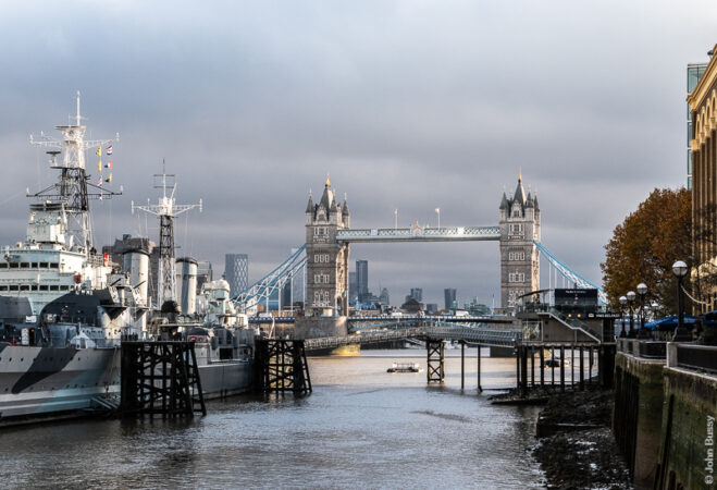 The sun is breaking through, under the clouds, onto Tower Bridge and HMS Belfast, with a reflection on the high-level walkway. (Nov25)